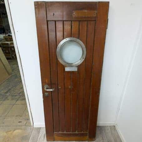 Full view of the salvaged ship's door with porthole window, highlighting the two-tone wood finish from exposure to elements.