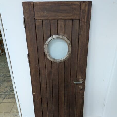 Angle view of a salvaged ship's door with a weathered porthole window and rustic hardware.